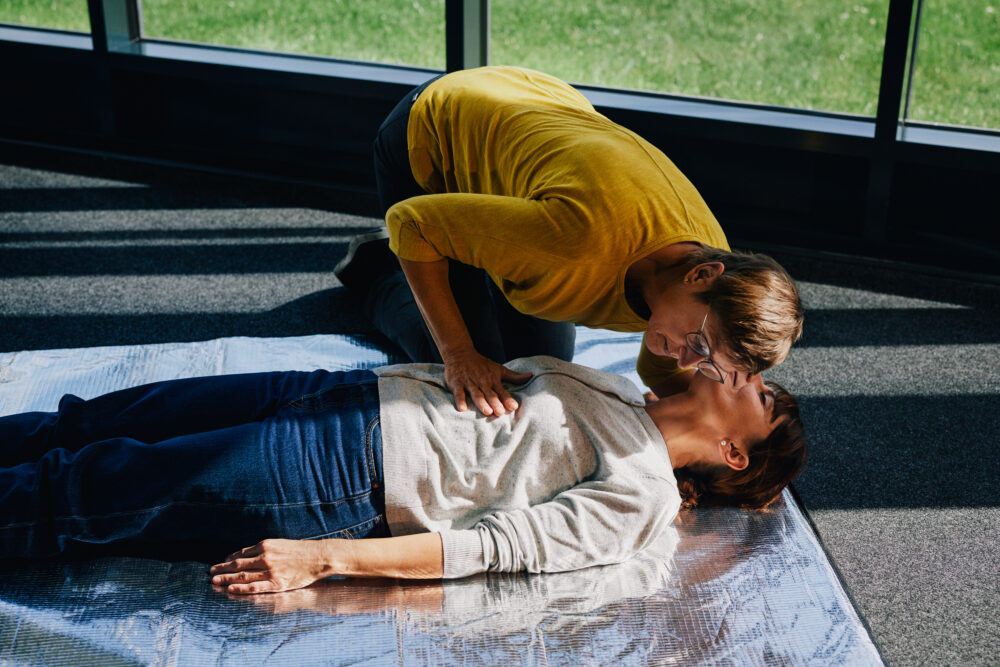 Frau stellt Erste-Hilfe-Übung auf dem Boden nach und horcht nach dem Atem der verletzten Person