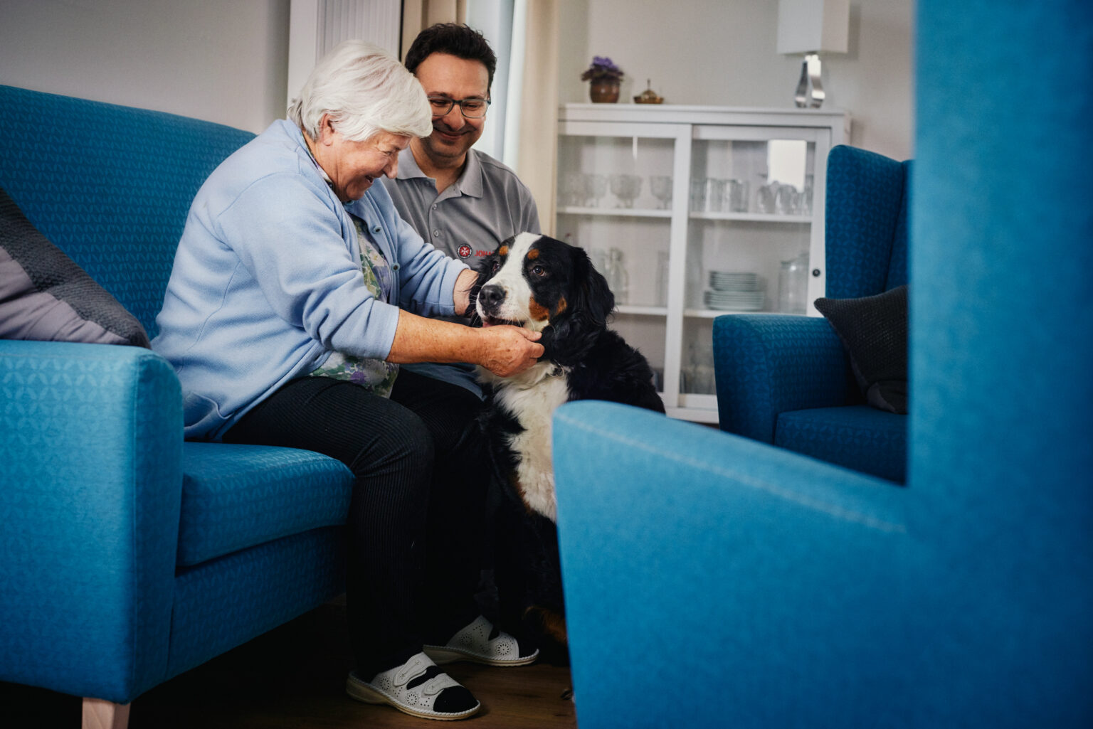 Ältere Dame und Pfleger sitzen gemeinsam auf der Couch und streicheln einen Hund