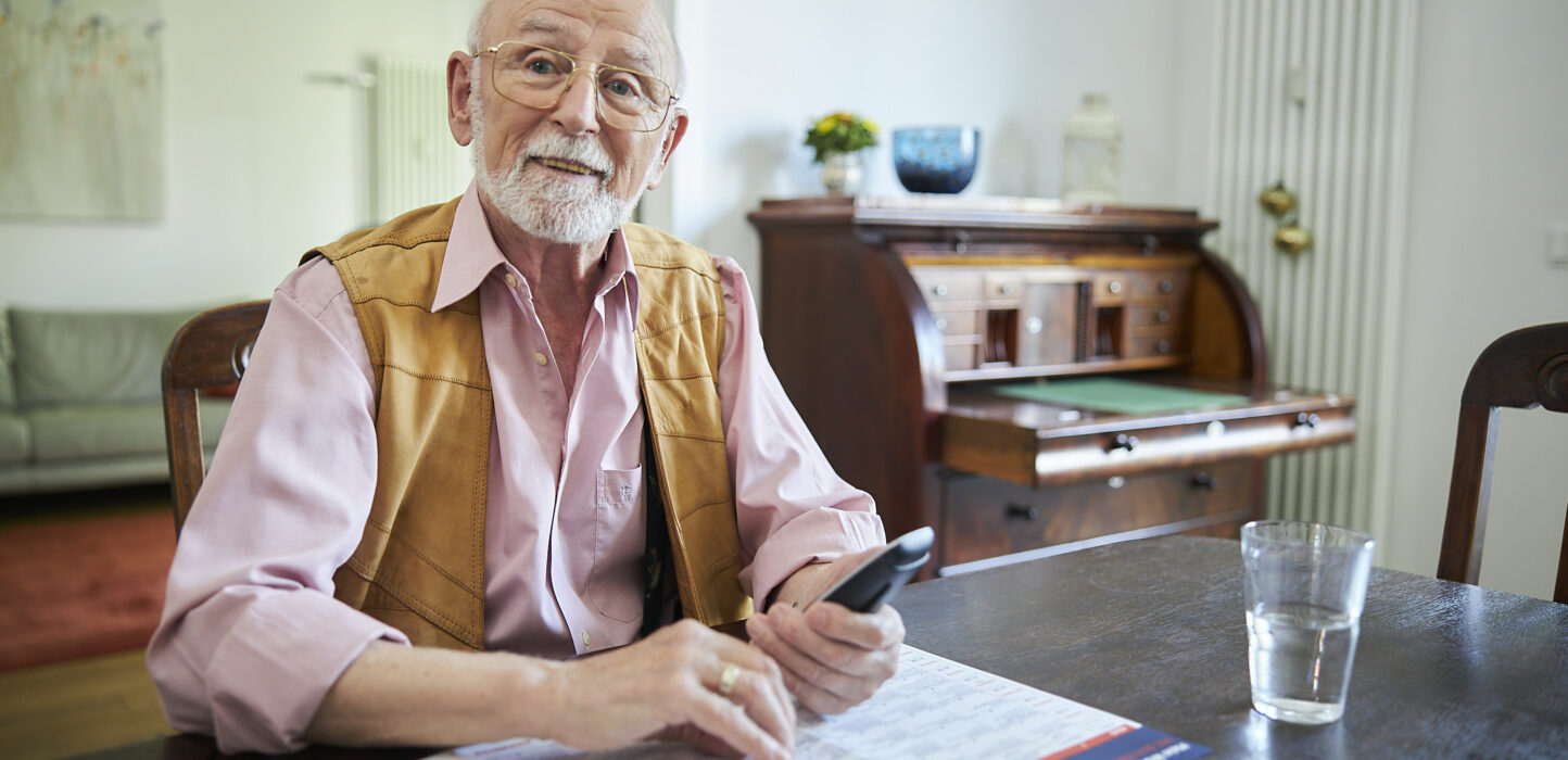 Älterer Mann hält ein Telefon in der Hand und vor ihm liegt eine aufgeschlagene Infobroschüre
