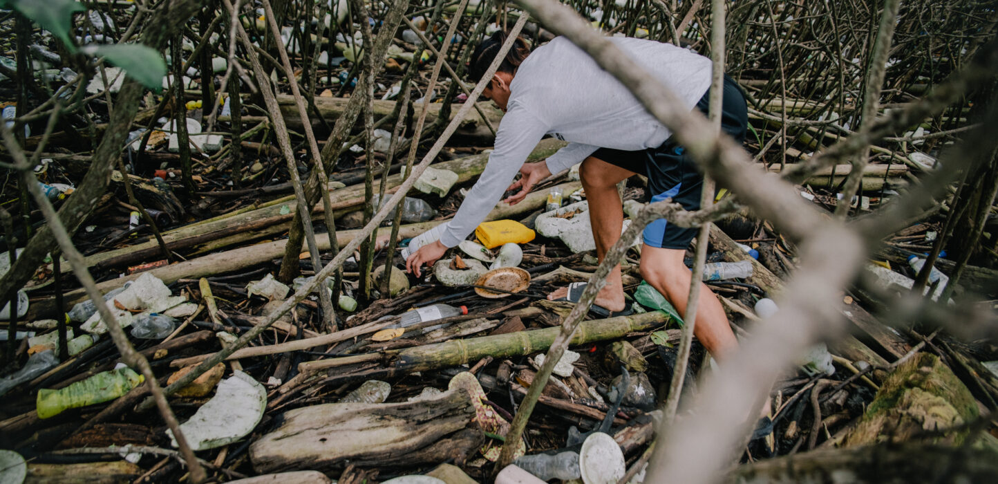 Mann sammelt Müll aus einem kleinen Waldstück, dass mit Ästen übersäht ist