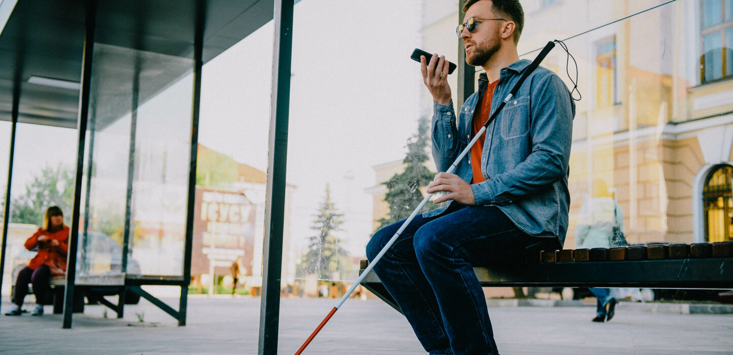 Junger blinder Mann mit Smartphone sitzt auf einer Bank in der Stadt und telefoniert