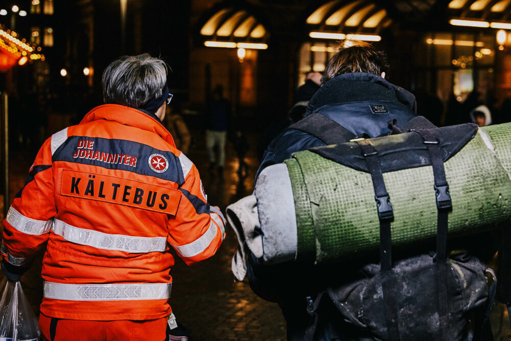 Frau mit orangener Arbeitsjacke vom Kältebus unterhält sich mit einem Obdachlosen
