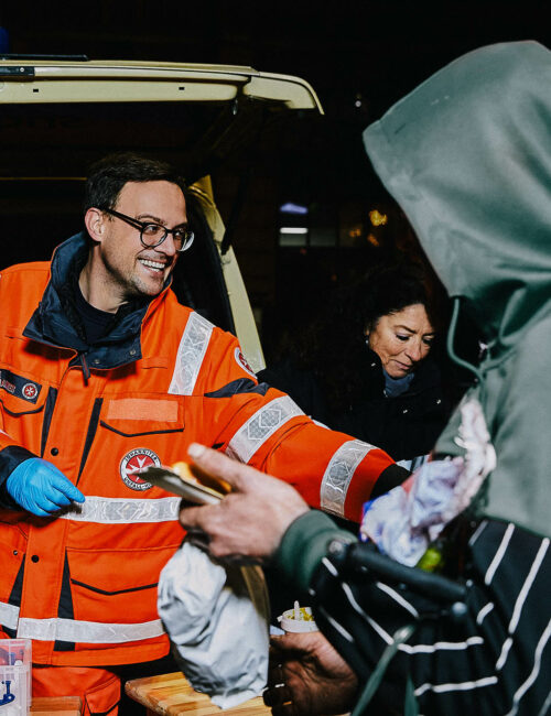 Mitarbeiter der JUH mit orangener Arbeitsjacke überreicht Essen an bedürftige Person