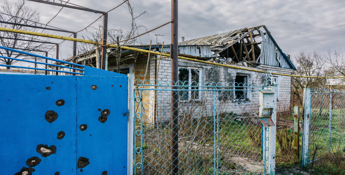 Ein baufälliges und zerstörtes Haus. Links ist eine blaue Wand mit Schusslöchern zu sehen.