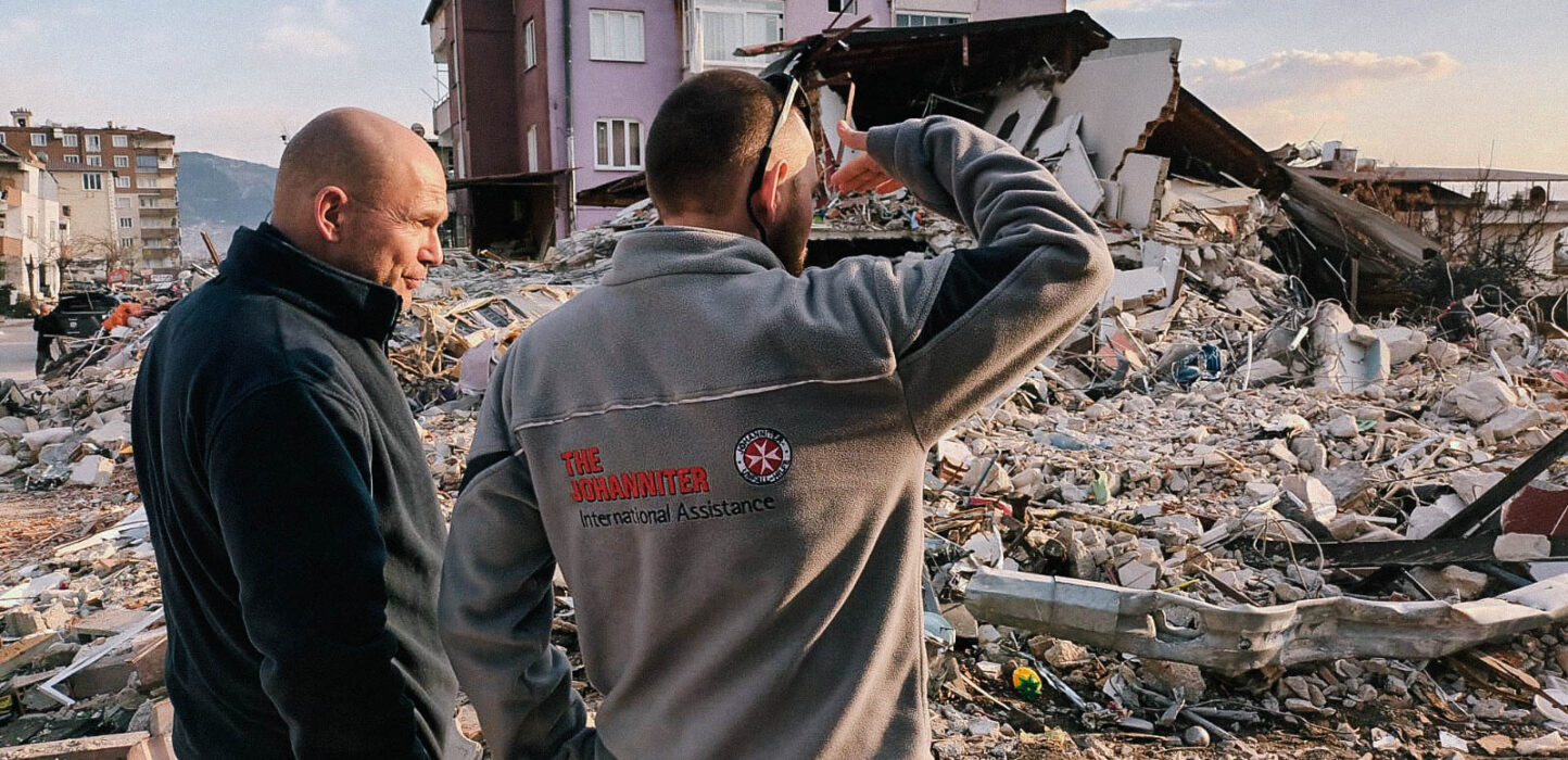 Zwei männliche Helfer der Johanniter stehen neben Trümmern und schauen auf die Erde.