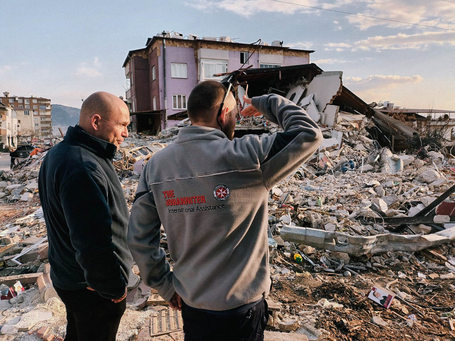 Zwei männliche Helfer der Johanniter stehen neben Trümmern und schauen auf die Erde.