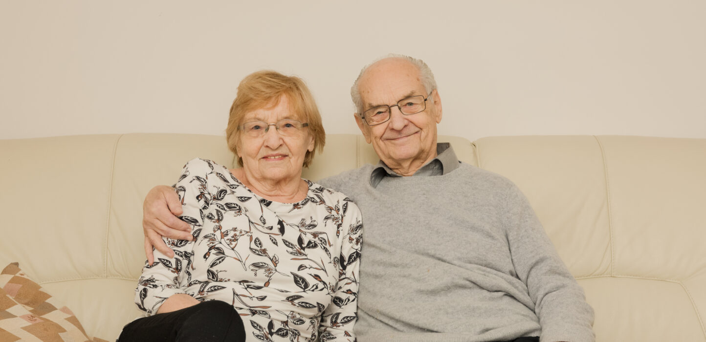 Älteres Ehepaar sitzt auf einer Couch und der Mann hat einen Arm um die Frau gelegt