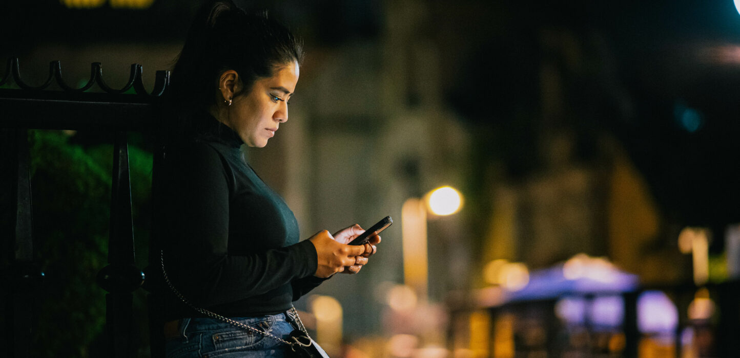 Frau sitzt alleine an einer beleuchteten Straße und schaut auf ihr Handy