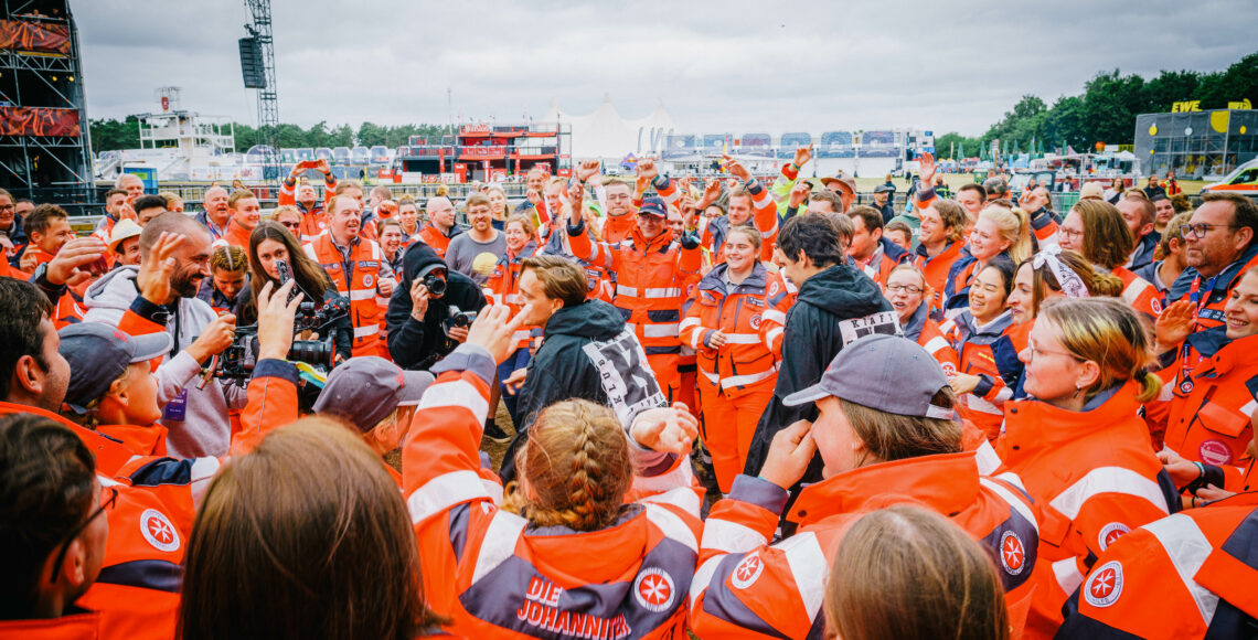 Eine Gruppe Rettungskräfte in orangen Uniformen stehen um Musiker in schwarzer Kleidung.
