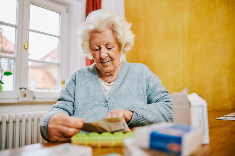 Ältere Frau mit hellblauem Cardigan sitzt am Tisch und schaut ihre Medikamente an.